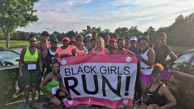 Before the start of their event, a group of runners pause on sunny morning in the park holding a Black Girls Run banner.