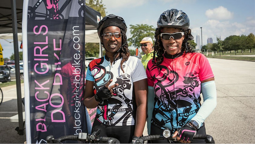 Two young black women smiling into the camera with helmets and biking gear on.