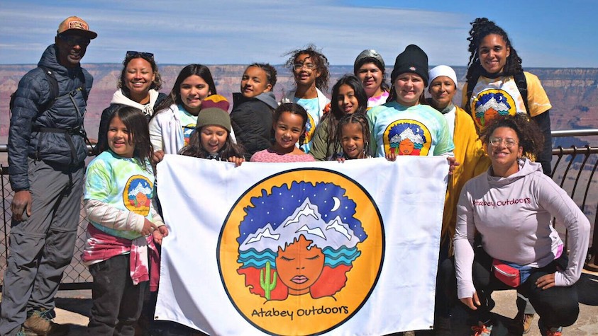 A diverse group of kids in the outdoors holding an Atabey Outdoors flag