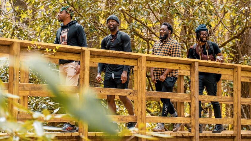 Four black men smiling as they cross a foot bridge while hiking in the woods. 