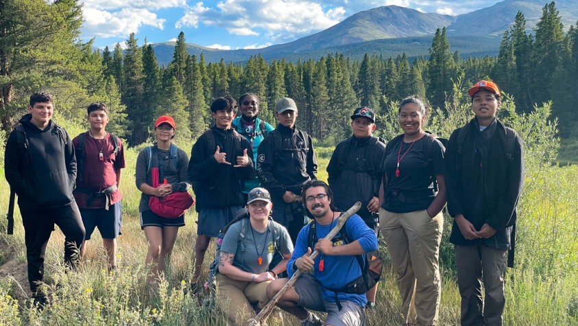 A diverse group of students smiling, standing in a field surrounded by a lush green hillside. 