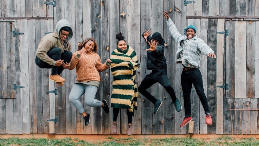 A group of five HBCU students dressed in warm clothing, jumping in front of a barn. 