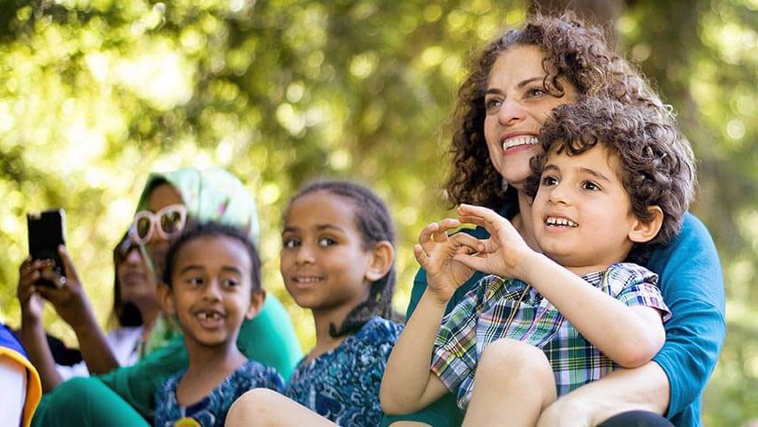 Kids and adults enjoy a sunny outdoor presentation under the trees.