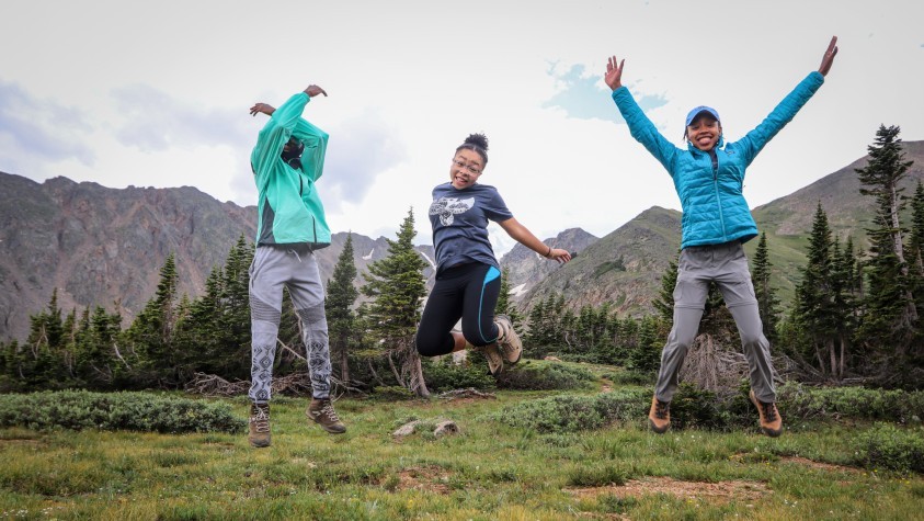 Three people jump in a field with mountains in the background