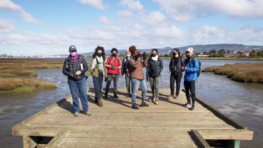 A group of people, wearing masks, stand at the end of a dock over the water