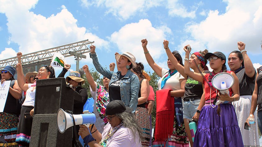 On stage, youths with megaphones raise their fists toward the sky at an outdoor gathering.