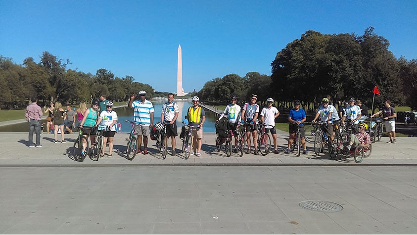 Framed by a bright blue sky punctuated by the Washington Monument, a group of cyclist pause for a photo op in front of the Lincoln Memorial Reflecting Pool.