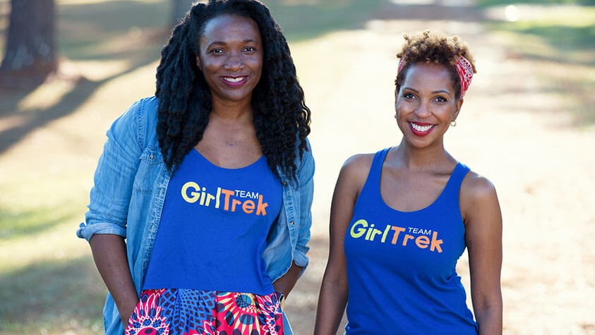 Two hikers wearing blue Team Girl Trek tee shirts smile for a photo.