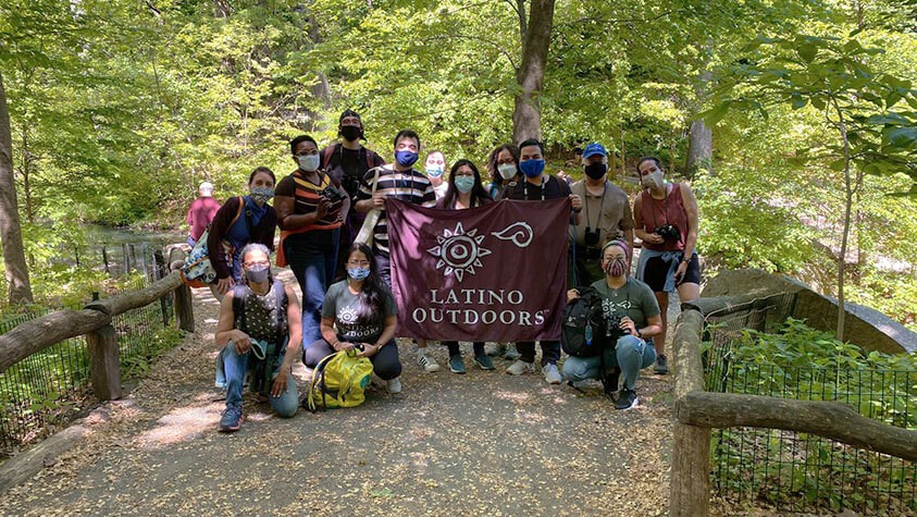 Wearing masks, a group of trail hikers holding a banner that reads Latino Outdoors pause for a afternoon photo op on a wooden bridge.