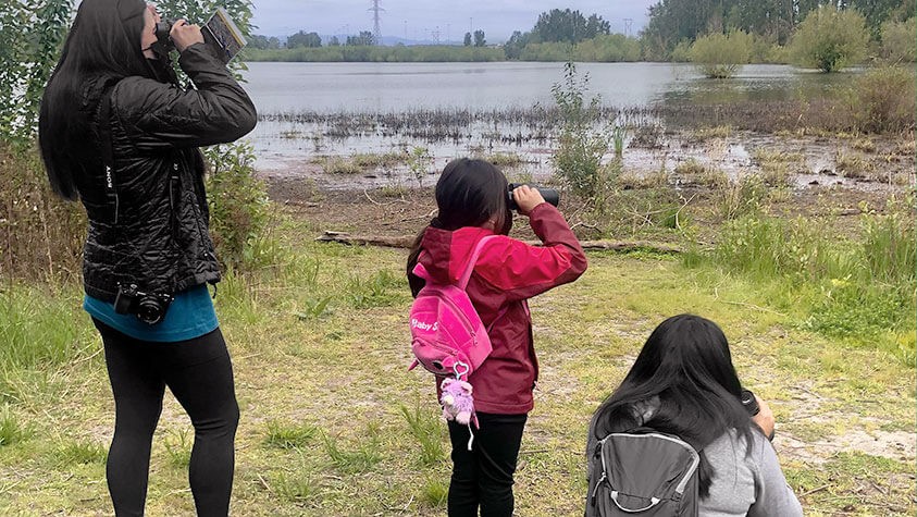 Using binoculars, three young hikers search for wildlife across a grassy lake.
