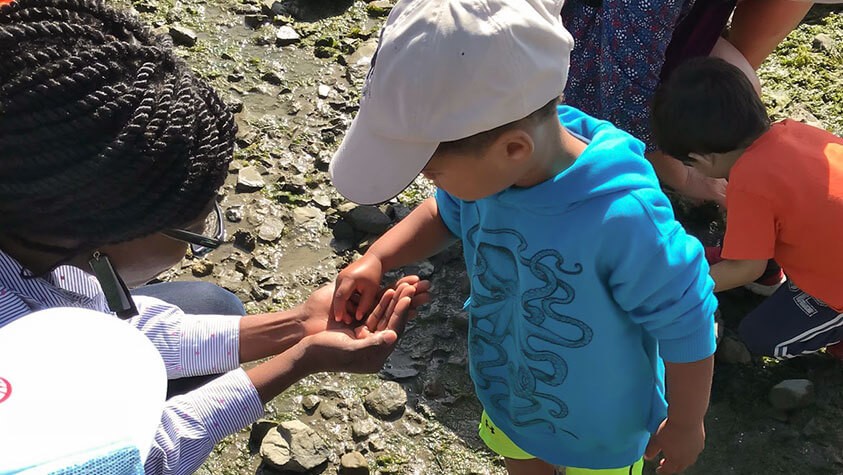 A parents helps their toddler, wearing a cap and blue octopus hoodie, explore the wonder of a tide pool.