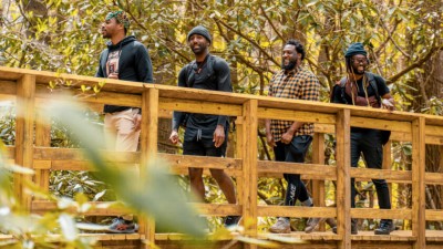 Four black men smiling as they cross a foot bridge while hiking in the woods.