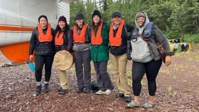 Six indigenous-identifying people stand smiling, shoulder-to-shoulder, under the wing of a small plane.