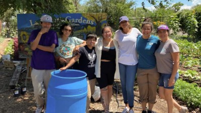 A group of seven diverse youth standing next to a rain barrel smiling in front of La Plazita Gardens. 