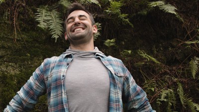 A person wearing a lightweight hoodie under a flannel shirt stands smiling with outstretched arms in front of a cluster of large ferns.