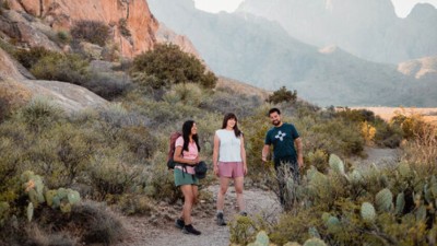Three people in short sleeves hiking a trail surrounded by cacti in front of a majestic New Mexico mountainside.
