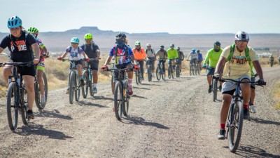 A large group of mountain biking youth taking a dusty path in front of an Arizona mountain range.