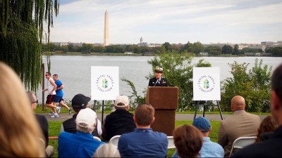Against a backdrop of the Washington Monument in the distance and framed by Capital Trails Coaltion signs, a Navy officer stands at a podium an addresses listeners.
