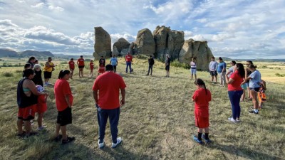 A large group of Native people standing in a circle under a partly cloudy Montana sky in front of large boulders.