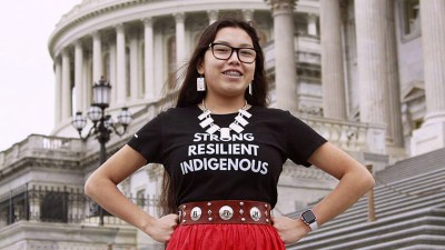 Hands on hip in front of the US capitol building, a youth wears a tee shirt that reads Strong, Resilient, Indigenous.