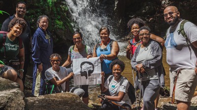 Pausing at the base of a waterfall, a group of happy hikers hold an Outdoor Afro banner.