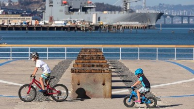 Against a backdrop of blue water and Navy vessels, two young bikers cruise the terrain of a sunny Bay area boardwalk, 