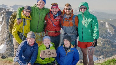 Under sunny skies, a group of alpine hikers stand at the summit holding a sign that reads Mount Ida, twelve thousand, eight hundred and twenty five feet.