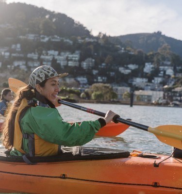  A group of three smiling kayakers paddle across Lake Washington in bright orange kayaks and yellow life vests, with the sun hitting their backs.