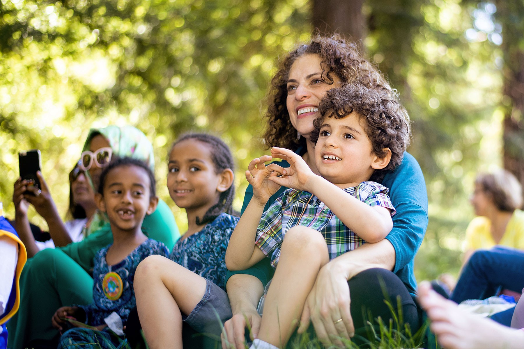 Kids and adults enjoy a sunny outdoor presentation under the trees.