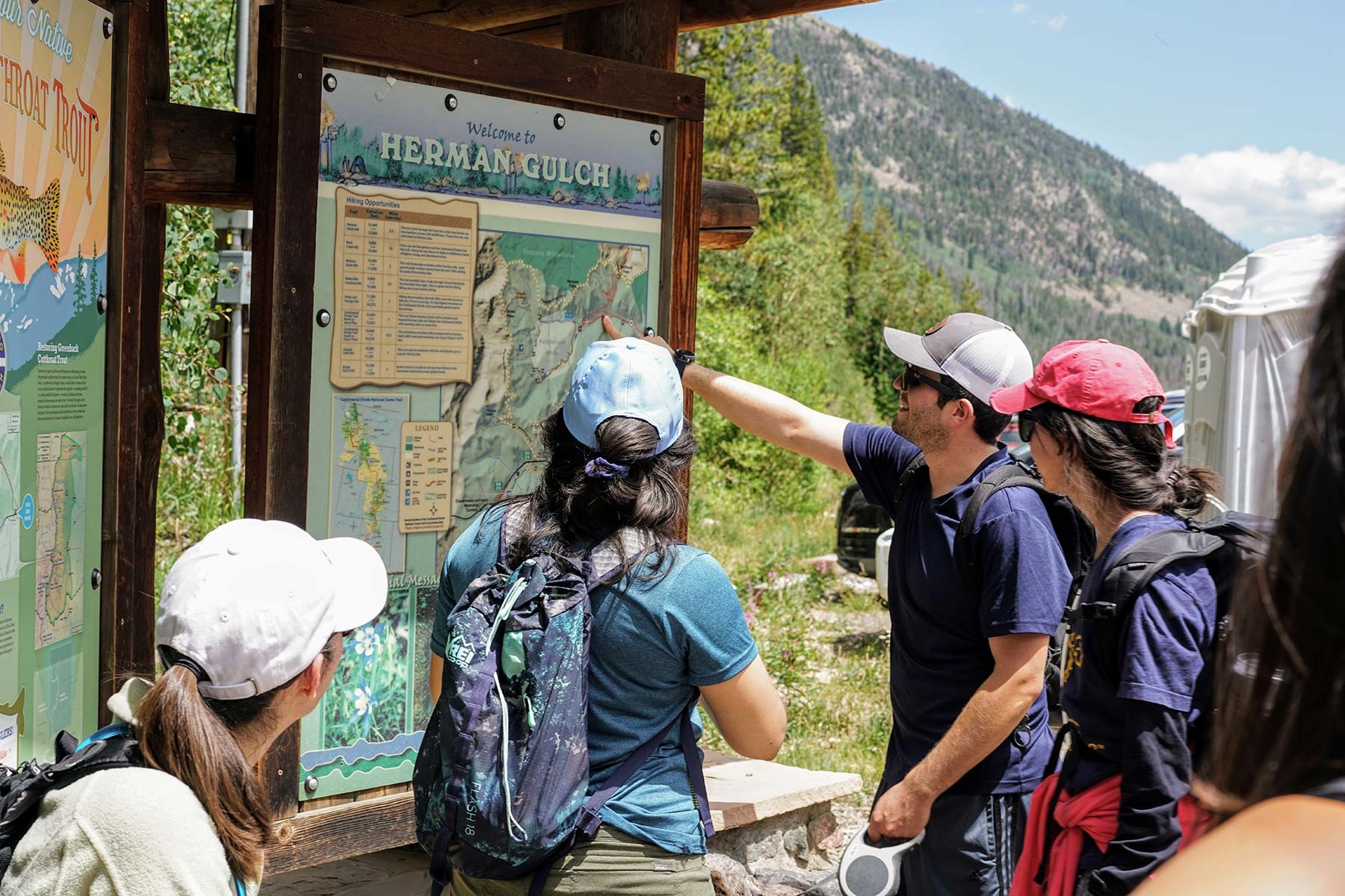 A group of four people wearing baseball caps look over a map of Herman Gulch, flanked by rocky hills. 