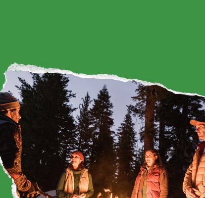 At dusk, four friends in puffy jackets stand around their campsite's fire, surrounded by fir trees.